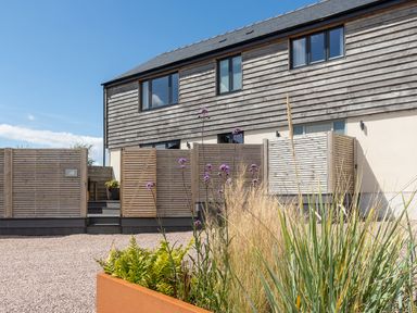 A modern two-story building with wooden cladding and a gravel courtyard with a planter of tall grasses and purple flowers at The Farmhouse in Llanfechell