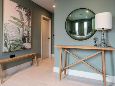 A hallway with a wooden bench a wooden console table with lamp and diffuser a large round mirror and botanical artwork at The Farmhouse in Llanfechell