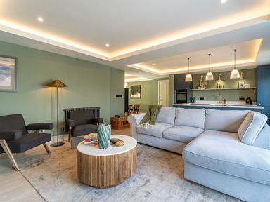 A living room with a light gray sectional sofa two black armchairs a round wooden coffee table and a kitchen area with pendant lights at The Farmhouse in Llanfechell