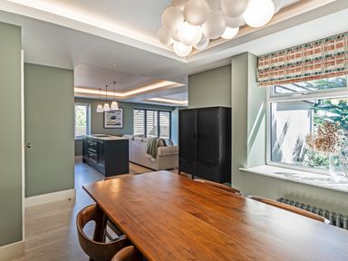 A dining area with a wooden table and chairs next to a window with a patterned blind and a black cabinet in an open plan living space at The Farmhouse in Llanfechell
