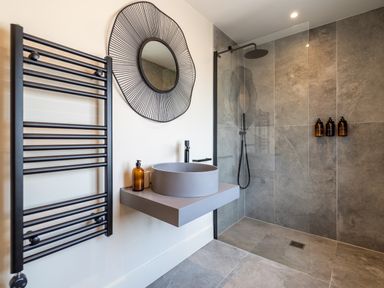 A bathroom with a round sink on a floating counter a round wall mirror a towel rack and a walk in shower with three bottles on the wall at The Farmhouse in Llanfechell