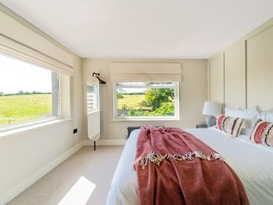 A bedroom with a bed covered with white sheets and red blankets and pillows next to two windows with a countryside view at The Farmhouse in Llanfechell