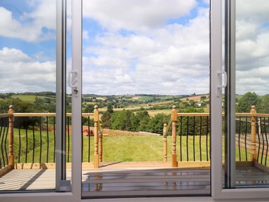 A view from sliding glass doors onto a wooden deck and a grassy yard with hills and fields in the distance at Ambleside in Cullompton