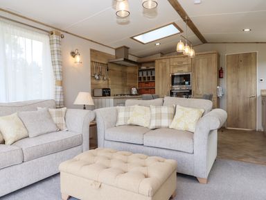 A living room with two sofas cushions and a padded ottoman in front of a kitchen with wooden cabinets at Ambleside in Cullompton