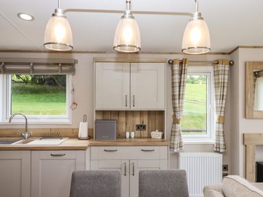 A kitchen with cabinets a sink a bread box paper towel holder and windows with curtains at Ambleside in Cullompton