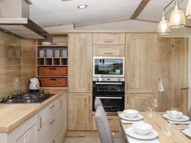 A kitchen with wooden cabinets a gas stove a kettle an oven and a dining table set with plates bowls and glasses at Ambleside in Cullompton