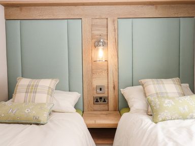 Two single beds with headboards and pillows and a wooden panel with a light and electrical outlets in between beds at Ambleside in Cullompton