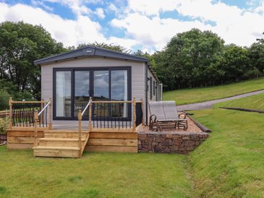 A small modular home with a wooden deck and steps facing a grassy area with two patio chairs on a stone platform at Ambleside in Cullompton