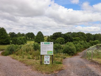 A gravel forked path with a rules sign in front of green trees under a cloudy sky at Ambleside in Cullompton