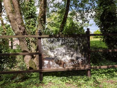 A wooden sign with a fish illustration and text Colebrook Fishery attached to a wooden fence surrounded by trees and greenery at Ambleside in Cullompton