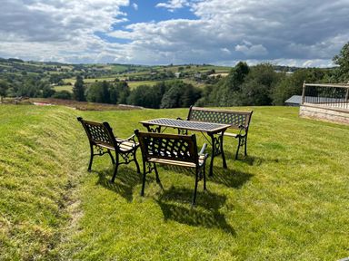 A metal and wood outdoor table with two chairs and a bench on a grassy hill with a rural landscape in the background at Ambleside in Cullompton