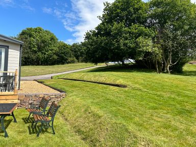 A grassy backyard with two chairs and a table next to a small building and large trees at Ambleside in Cullompton
