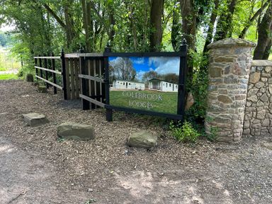 A wooden fence and stone pillar with a Colebrook Lodges sign beside a wooded area at Ambleside in Cullompton