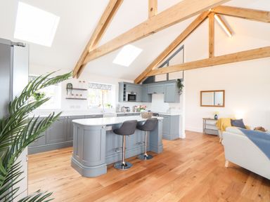 A kitchen with blue cabinets and a white countertop island with two stools next to a living area at Trevilges Cottage in Trewennack near Helston