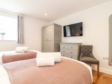 A bedroom with two beds with towels on them a grey wardrobe a dresser a chair and a wall-mounted television at Trevilges Cottage in Trewennack near Helston