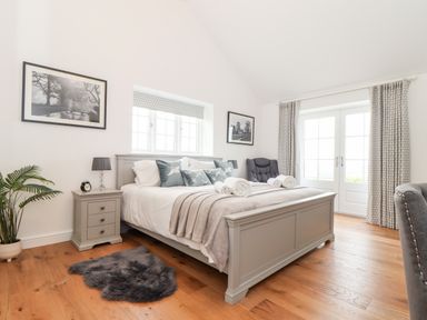 A bedroom with a gray bed frame and bed with pillows and towels two nightstands with lamps a gray rug wooden floor and doors with curtains at Trevilges Cottage in Trewennack near Helston