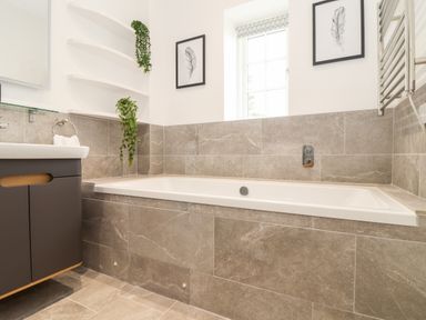 A bathroom with a bathtub surrounded by gray tiles a window above the bathtub a sink with cabinet on the left and framed pictures on the wall at Trevilges Cottage in Trewennack near Helston