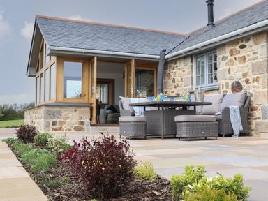An outdoor patio with a round table and cushioned chairs next to a stone house with wooden framed glass doors and windows at Trevilges Cottage Trewennack near Helston