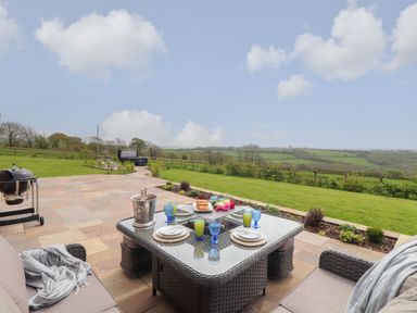 An outdoor patio area with a wicker dining table set with plates and glasses a grassy garden and countryside view at Trevilges Cottage in Trewennack near Helston