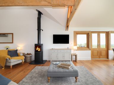 A living room with a wood burning stove a gray ottoman a wooden floor and wooden framed windows at Trevilges Cottage in Trewennack near Helston