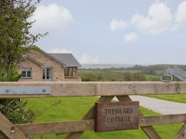 A wooden gate with a sign reading Trevilges Cottage with a stone and wood house and green fields in the background at Trevilges Cottage Trewennack near Helston
