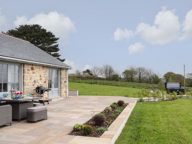 An outdoor patio area with a dining table and chairs a barbecue and a garden with lawn and shrubs at Trevilges Cottage Trewennack near Helston