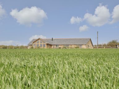 A stone house with large windows behind a fenced green field under a blue sky at Trevilges Cottage in Trewennack near Helston