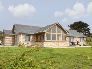A single-story stone house with large wooden framed windows and a sloped roof surrounded by grass at Trevilges Cottage in Trewennack near Helston