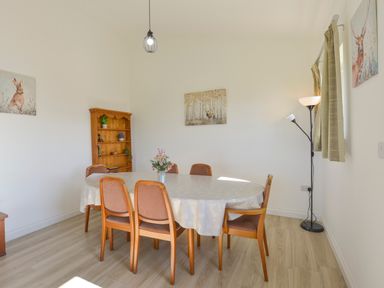 A dining room with an oval table covered with a tablecloth surrounded by six chairs and a wooden shelf with plants at The Linhay in Lapford