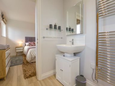 A bathroom sink with a mirror and toiletries and a partial view of a bedroom with a bed and dresser at The Linhay in Lapford