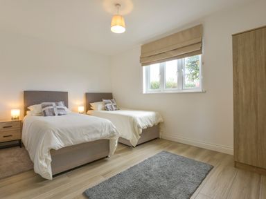 A bedroom with two single beds a bedside table with lamps a window with blinds a gray rug and a wooden wardrobe at The Linhay in Lapford
