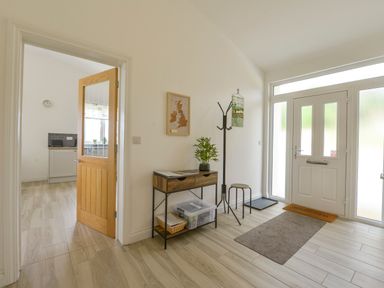 A hallway with a wooden door leading to a kitchen a console table with a plant a coat rack a stool and a door with frosted glass panels at The Linhay in Lapford