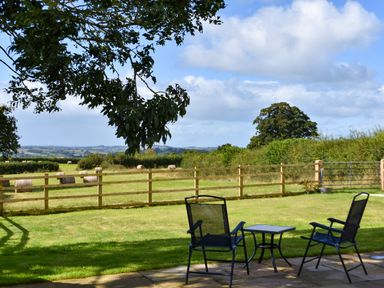 Two chairs and a small table on a patio overlooking a fenced grassy field with hay bales and trees at The Linhay in Lapford