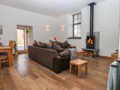 A living room with a brown sofa, wooden stools, a wood-burning stove, a dining table, and a window at The Old Chapel in Ilsington