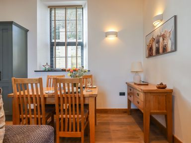 A dining area with wooden table and chairs flowers on table sideboard with lamp and bowl painting on wall at The Old Chapel in Ilsington