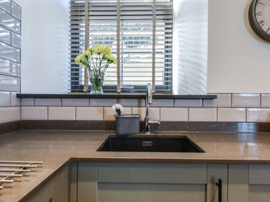 A kitchen sink with a faucet and a dish brush on the countertop near a window with blinds and flowers on the windowsill at The Old Chapel in Ilsington