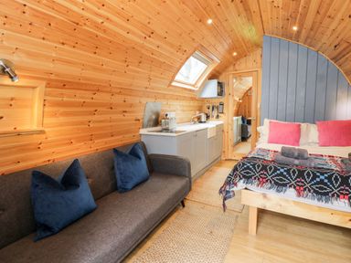 A wooden pod interior with a gray sofa and blue pillows a bed with pink pillows and folded towels a kitchenette with appliances and a skylight window at Conwy Pod in Capel Garmon near Betws-Y-Coed