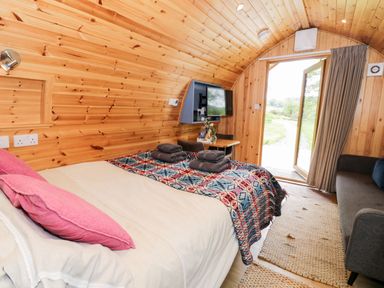 A wooden interior bedroom with a bed, pink pillows, folded towels, a small table with chairs, a television, and an open door showing an outdoor path at Conwy Pod in Capel Garmon near Betws-Y-Coed