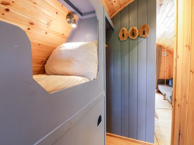 A sleeping pod with a pillow and light in a wooden cabin hallway with three wooden wall hooks and a glimpse of a sofa at Conwy Pod in Capel Garmon near Betws-Y-Coed