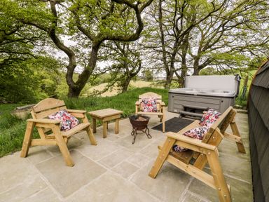 An outdoor patio with wooden chairs and floral cushions around a fire pit and a hot tub at Conwy Pod in Capel Garmon near Betws-Y-Coed