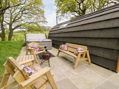 An outdoor patio with wooden chairs and benches with floral cushions a fire pit and a hot tub next to a pod with black shingles Conwy Pod Capel Garmon near Betws-Y-Coed