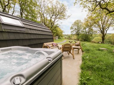 An outdoor patio area with a hot tub wooden benches chairs and a picnic table surrounded by grass and trees at Conwy Pod in Capel Garmon near Betws-Y-Coed