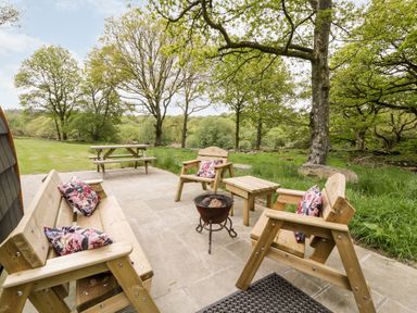 An outdoor patio area with wooden benches and chairs around a small fire pit and a picnic table in a grassy area with trees at Conwy Pod in Capel Garmon near Betws-Y-Coed