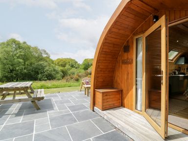 A wooden curved cabin with an open door on a stone patio with a wooden picnic table and benches outside Llugwy Pod Capel Garmon near Betws-Y-Coed