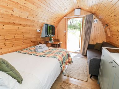 A wooden pod interior with a bed, a sofa, a table with chairs, a TV, and an open door at Llugwy Pod in Capel Garmon near Betws-Y-Coed