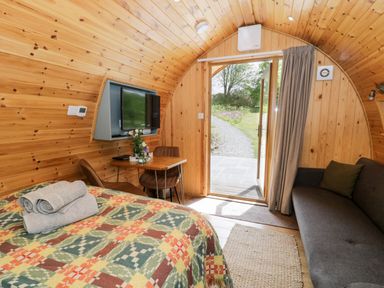 A wooden pod interior with a bed covered with a patterned blanket and towels a small table with two chairs a wall-mounted television a sofa and an open door leading outside at Llugwy Pod Capel Garmon near Betws-Y-Coed