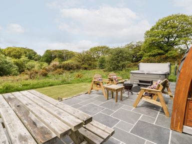 An outdoor patio with wooden chairs a table a fire pit a hot tub and a picnic table surrounded by grass and trees at Llugwy Pod in Capel Garmon near Betws-Y-Coed