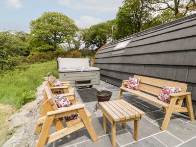 An outdoor patio with wooden chairs and a bench with floral cushions a small table a fire pit and a hot tub beside a curved building at Llugwy Pod in Capel Garmon near Betws-Y-Coed