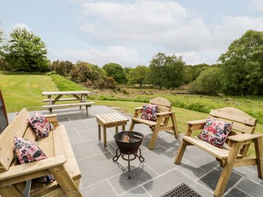 An outdoor patio with wooden benches and chairs with floral cushions a small table and a barbecue grill with grass and trees in the background at Llugwy Pod in Capel Garmon near Betws-Y-Coed