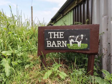 A wooden sign with a painting of a cow and the words the barn in tall grass beside a metal building at Northwood Barn in Okehampton
